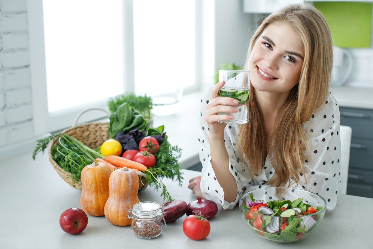 Woman holding glass of water surrounded by fresh vegetables and salad in bright kitchen.