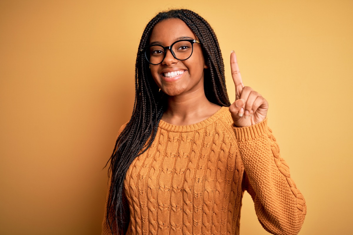 Young woman with glasses wearing orange sweater, smiling and pointing upward against yellow background.