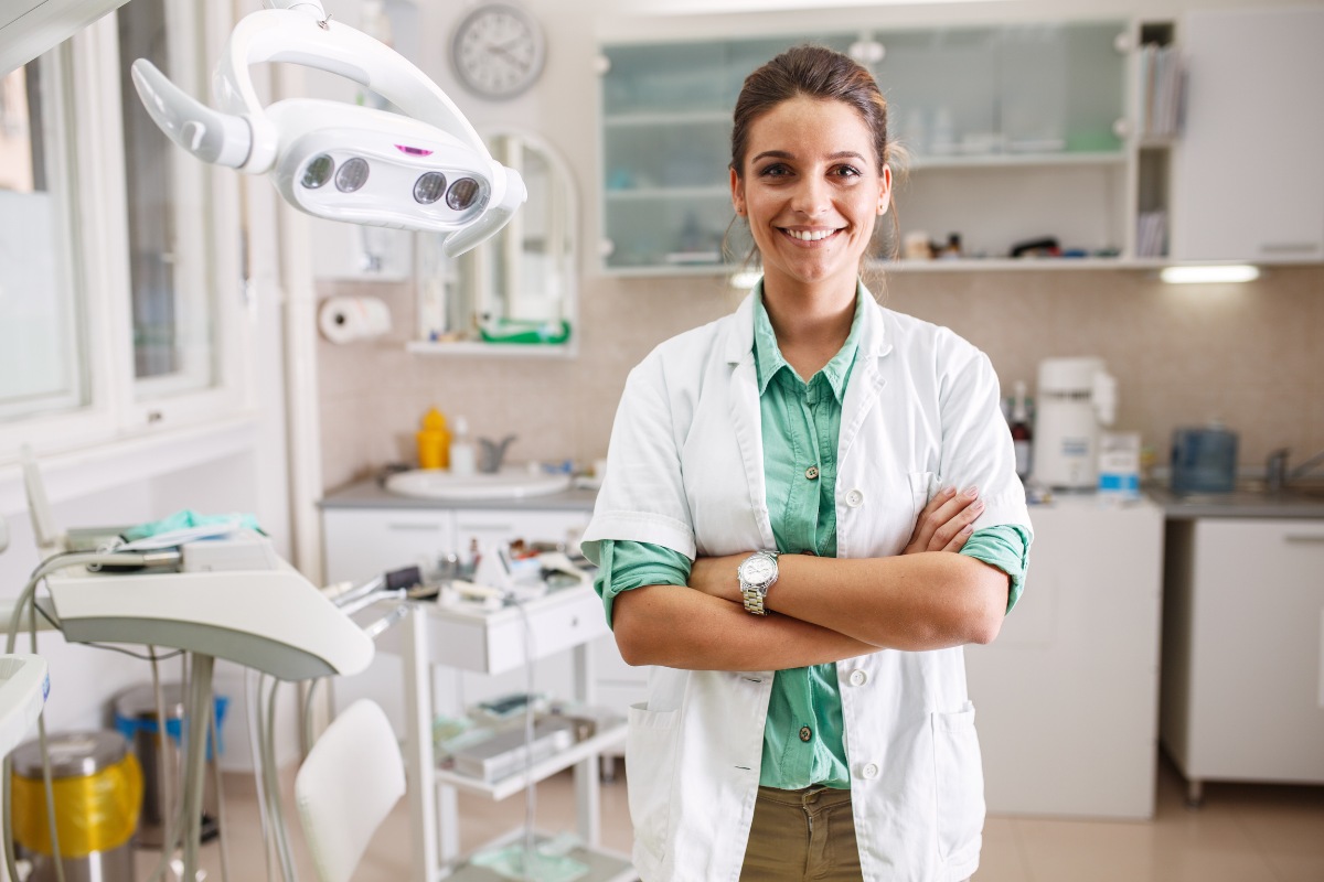 Female dentist in white coat standing with crossed arms in modern dental clinic office.