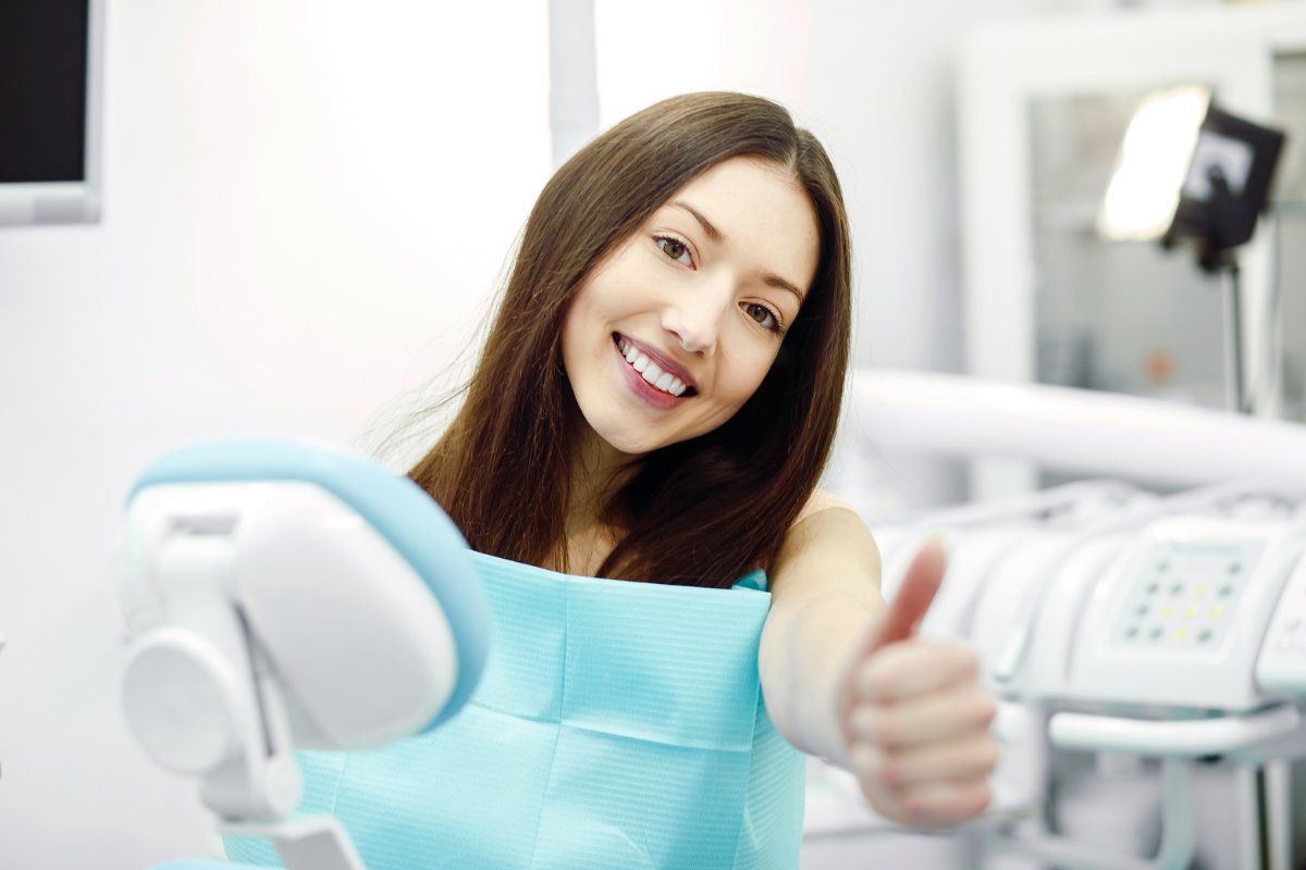 Woman in dental chair giving thumbs up, smiling at dentist office with equipment visible.