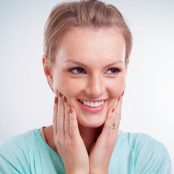 Woman in turquoise shirt smiling with hands on face against white background.