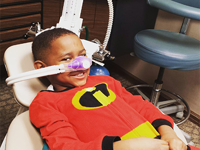 Young child wearing red Incredibles shirt in dental chair with purple glasses during exam.