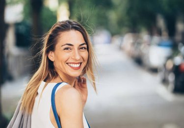 Woman smiling at camera on urban street with blurred background and parked cars.
