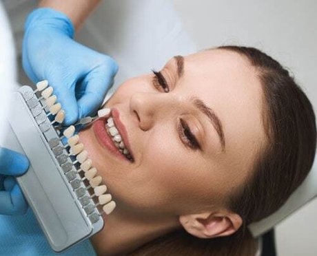 Dental professional holding tooth shade guide next to patient's smiling face during color matching consultation.