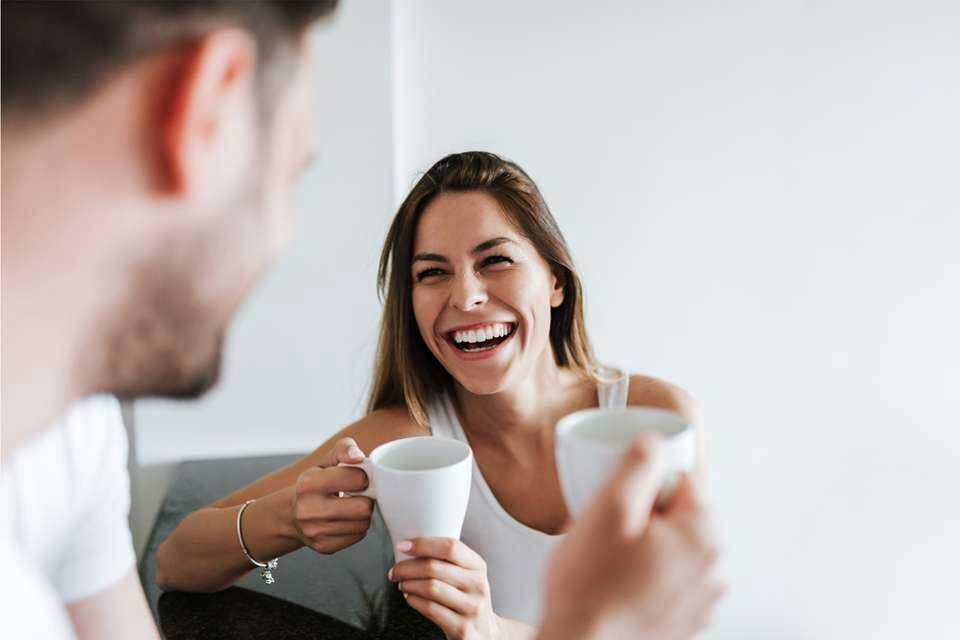 Two people smiling while holding coffee cups in a bright, casual indoor setting.