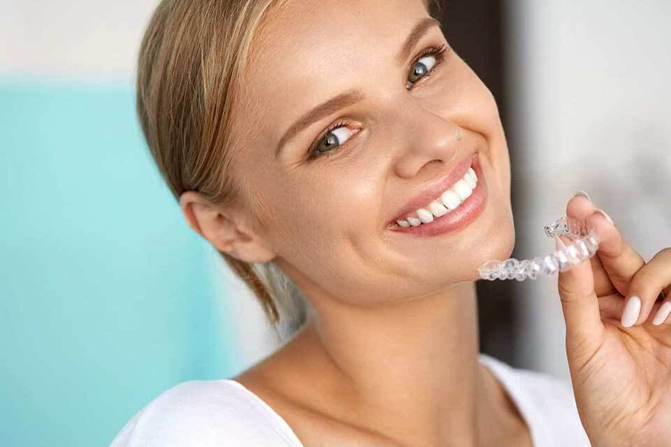 Woman brushing teeth with electric toothbrush against blue background, smiling at camera.