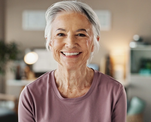 img-who-is-a-candidate-for-dental-implants Smiling older woman with gray hair wearing mauve shirt in bright home interior